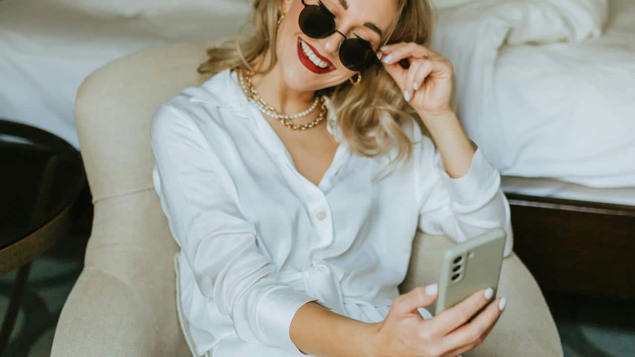 A smiling woman in a white blouse taking a selfie with her smartphone.