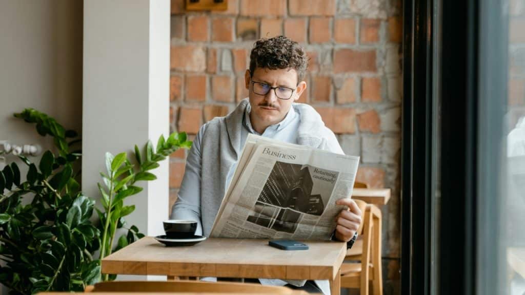 A man with glasses sits alone at a cafe table reading a business newspaper.