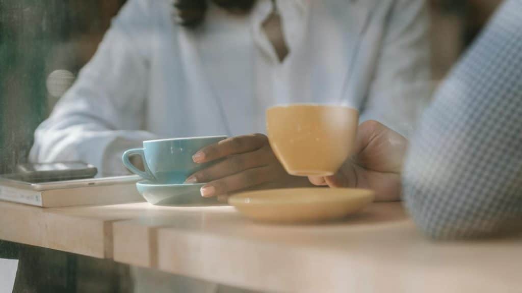 A person holding a coffee cup while sitting at a café table with another cup and a phone nearby.