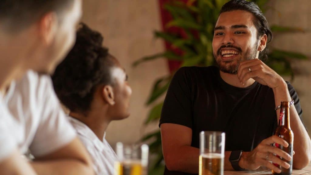 A man smiling and talking with friends while holding a beer at a table.