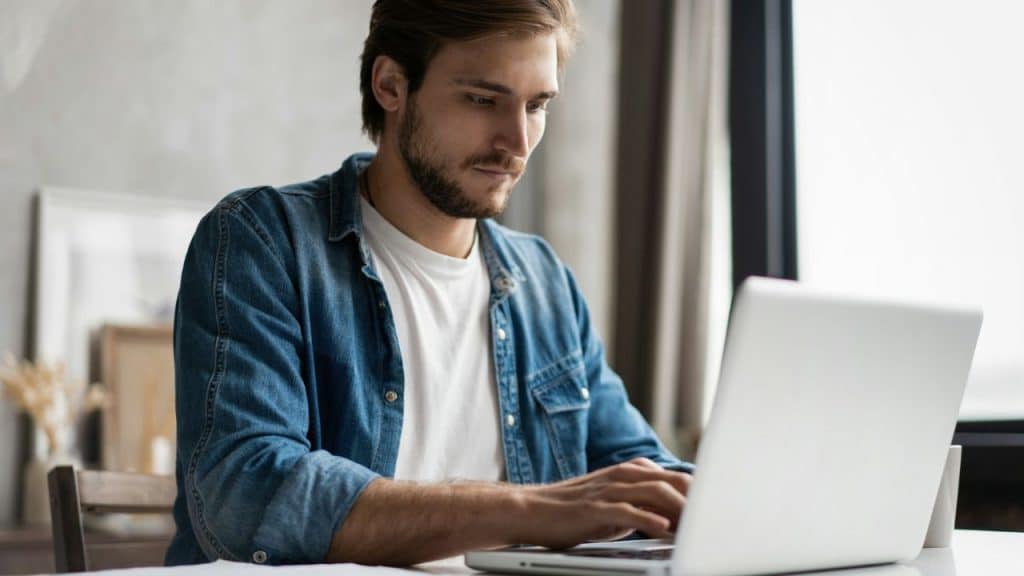 A man with a beard wearing a denim shirt over a white shirt uses a laptop.