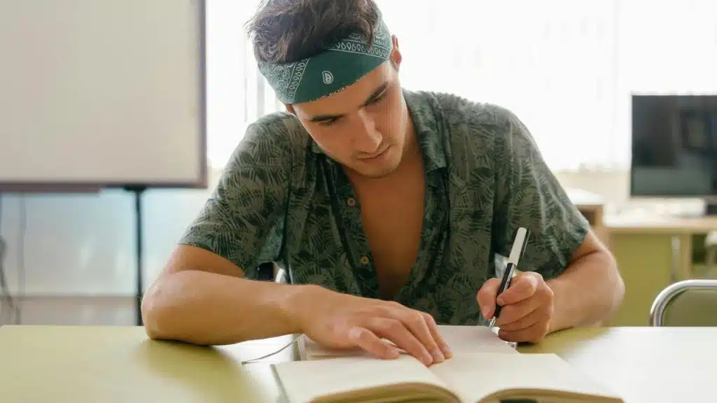 A young man wearing a bandana writing in a notebook at a desk.