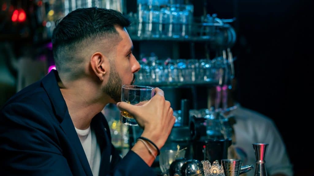 A man in a dark jacket sits at a bar holding a glass of liquid.