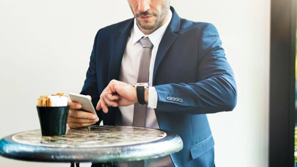 A man in a suit looks at his wristwatch while holding a smartphone.