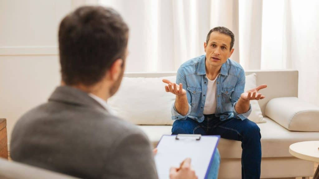 A man sits on a couch talking to a professional holding a clipboard.