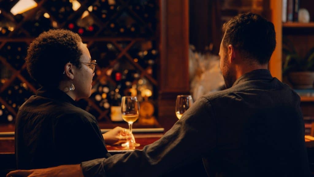 A couple sits at a bar holding wine glasses while facing away from camera.