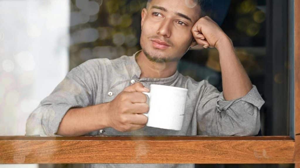 A man holding a mug and looking thoughtfully out a window.
