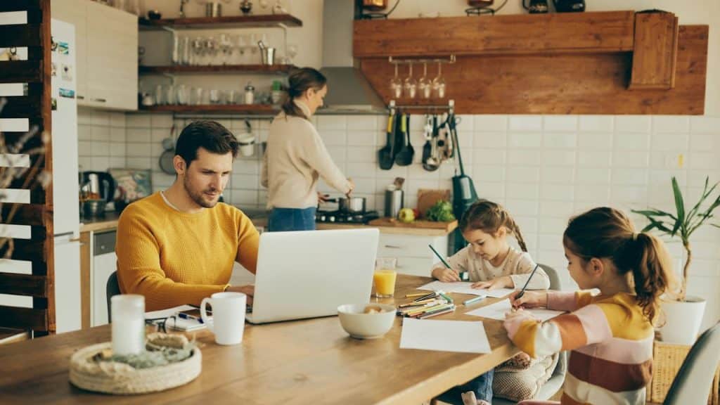 A man works on a laptop at a table while two children draw beside him.
