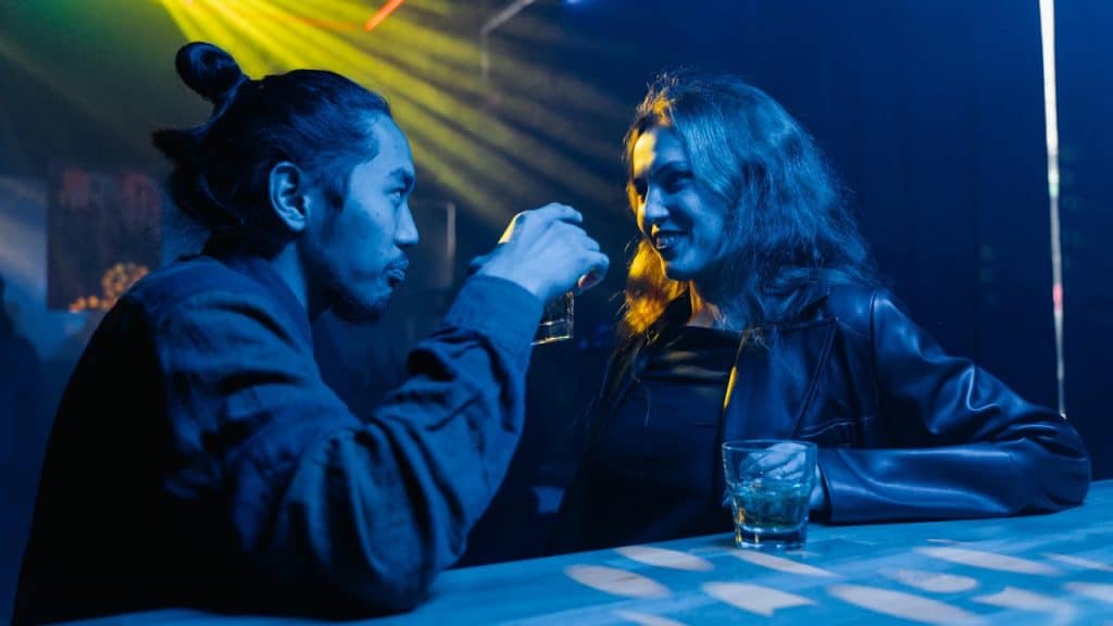 A man and woman sit at a dark bar under blue and yellow lights with drinks.