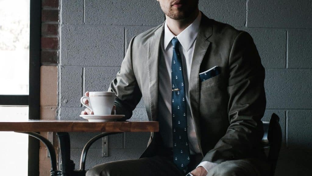 A man wearing a suit and tie sits at a table holding a cup.