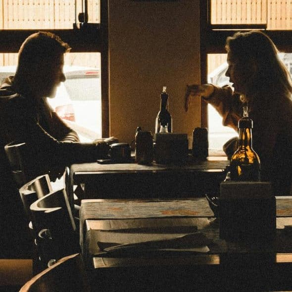 A backlit café scene with two people talking across a table by a window.