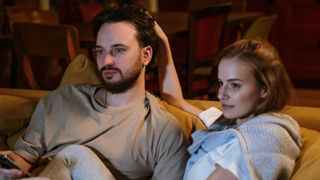 A man and woman sit closely on a couch while looking toward a screen.