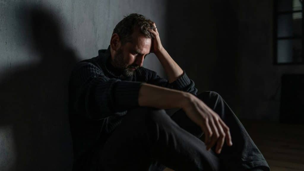 A man in a sweater sits on the floor in shadows, holding his head.