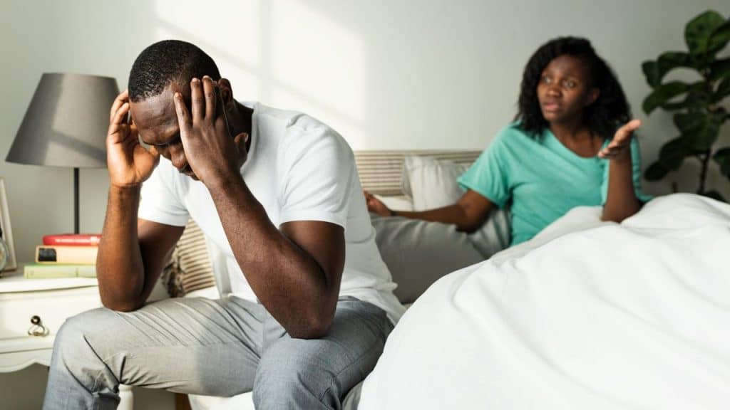 A man sits on a bed holding his head while a woman gestures nearby.