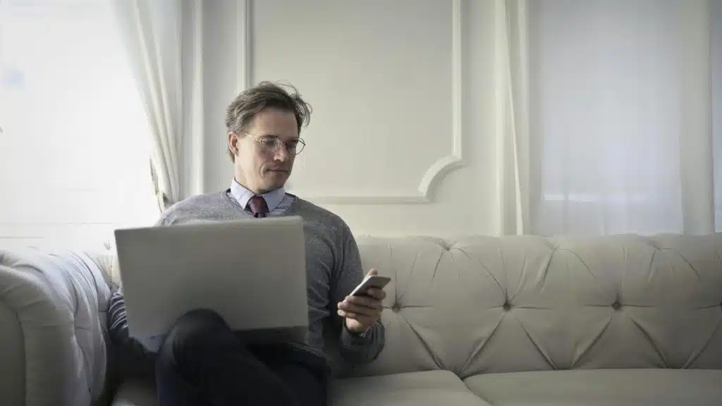 A man sitting on a couch using a laptop and phone.