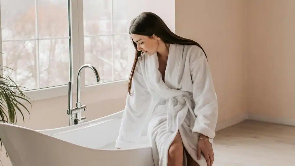 A woman in a white bathrobe tests the water in a bathtub.