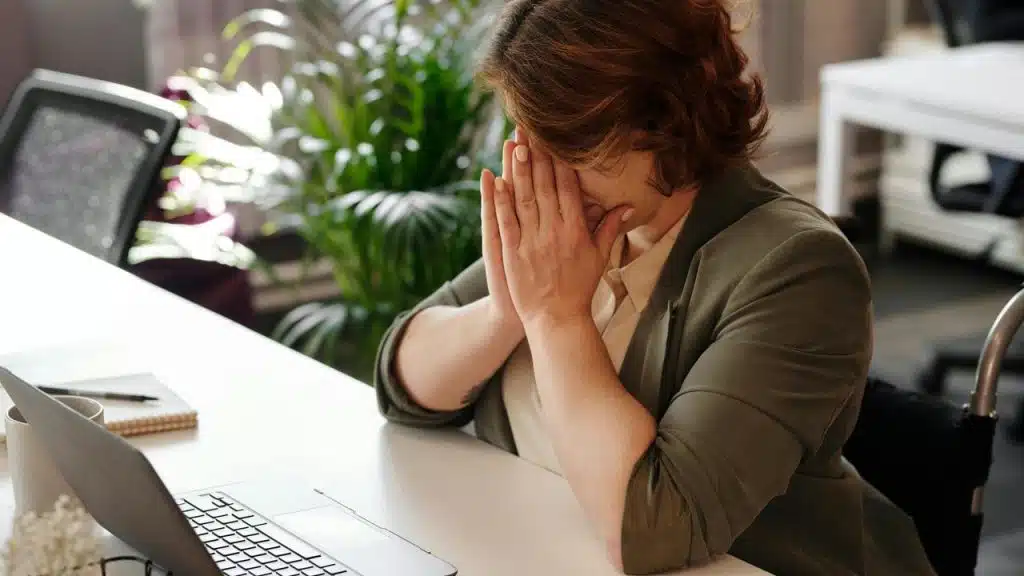 A stressed person covering their face while sitting at a desk with a laptop.