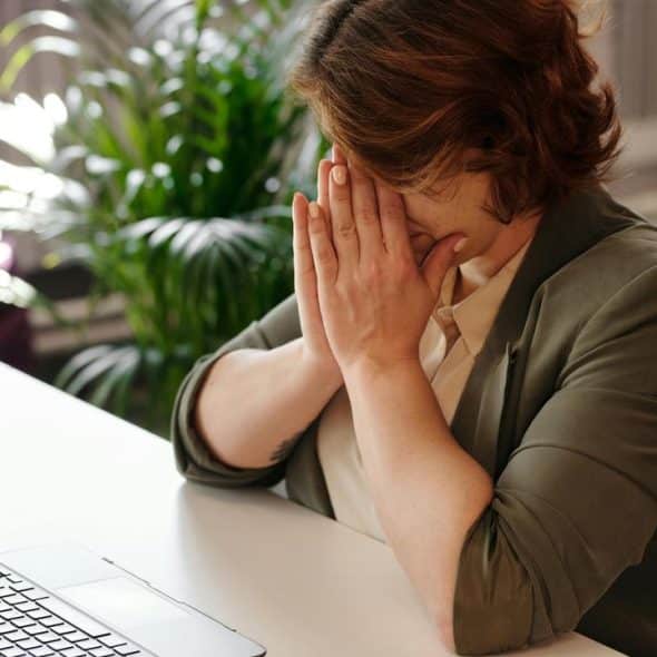 A stressed person covering their face while sitting at a desk with a laptop.