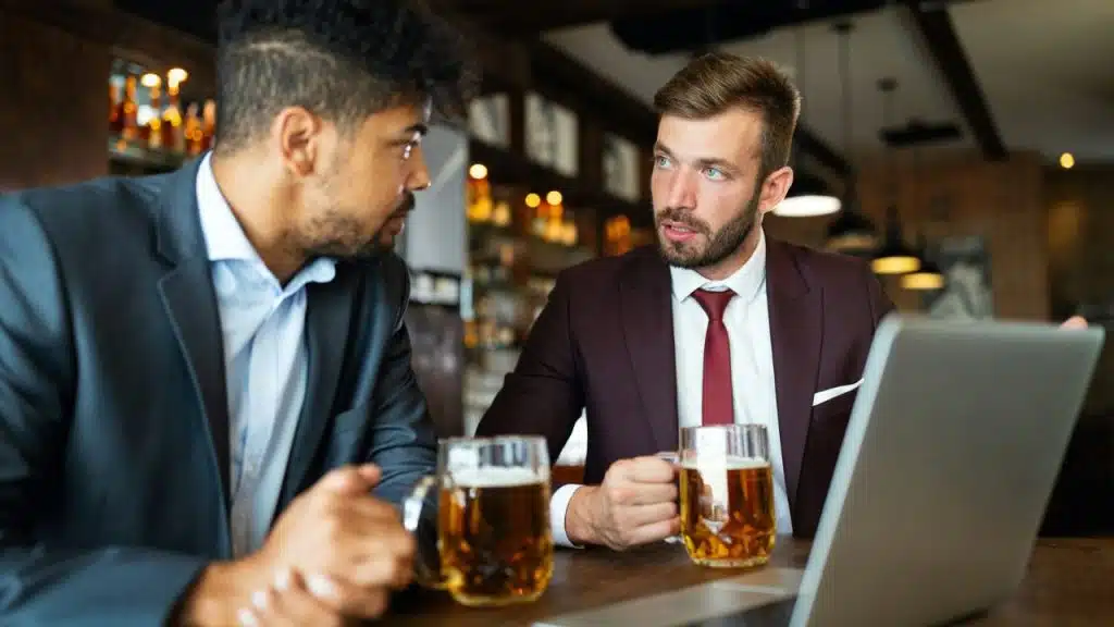 Two men in suits sit at a bar with mugs, seriously talking.