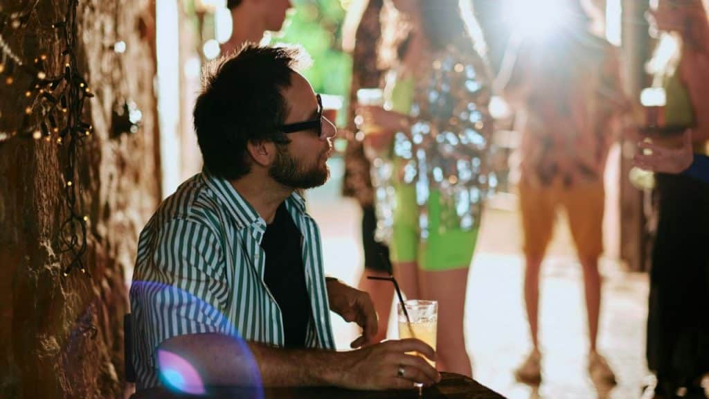 A man in a striped shirt holds a drink near a blurred group gathering.