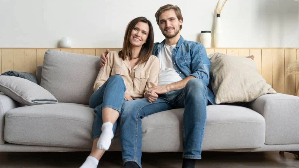 A man and woman sit together on a gray sofa smiling at the camera.
