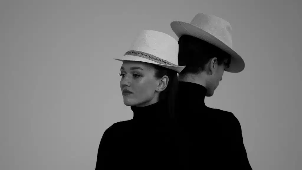 Two people in black turtlenecks and wide-brimmed hats standing back to back.