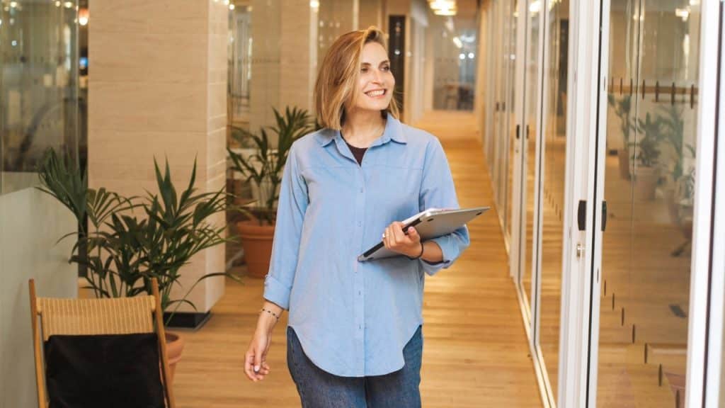 A smiling woman walks down a bright hallway carrying a laptop and a notebook in hand.