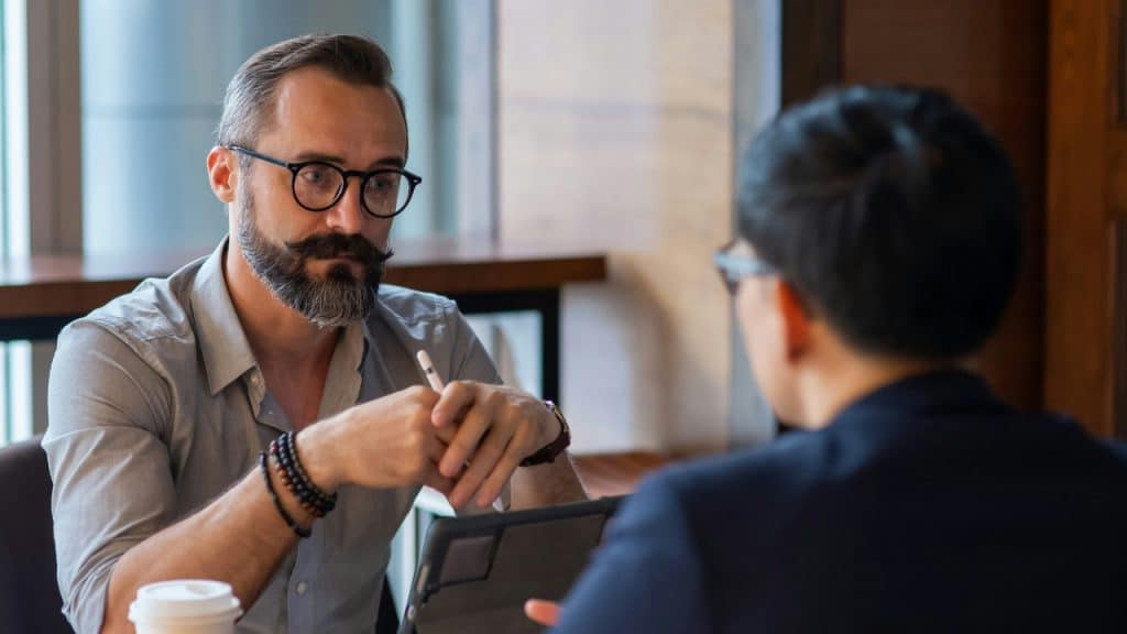 A man with a mustache and glasses looks at another person while holding a pen.