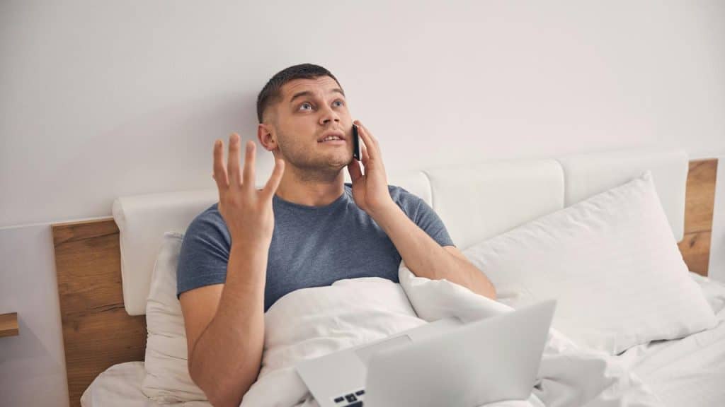 A man sitting in bed talking on the phone with a laptop in front of him.