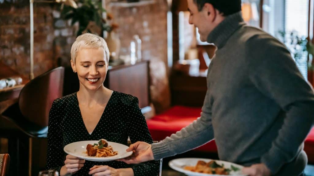 A man hands a plate of food to a smiling woman at a restaurant table.