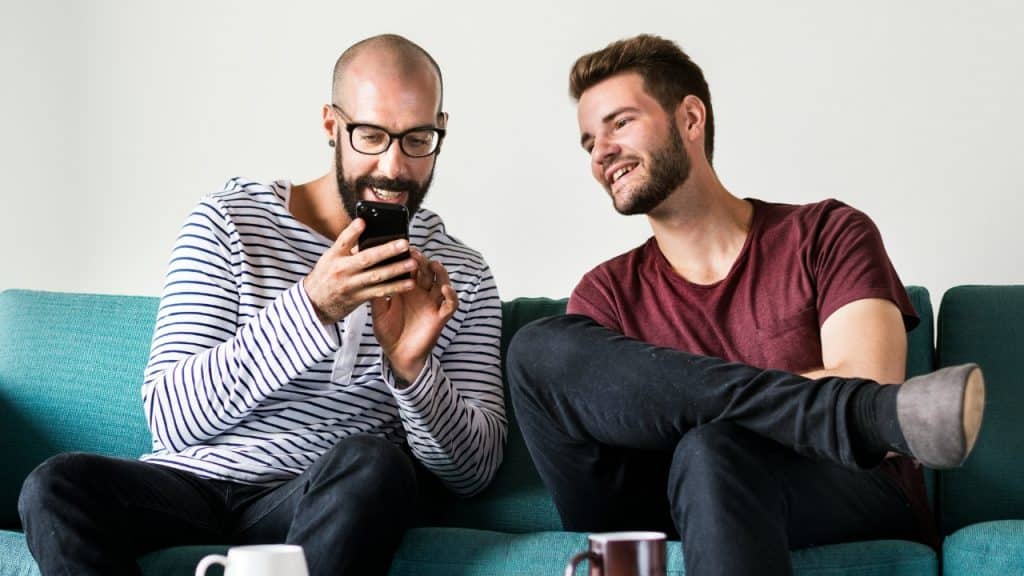 Two men sit on a green sofa looking at a smartphone held by one.