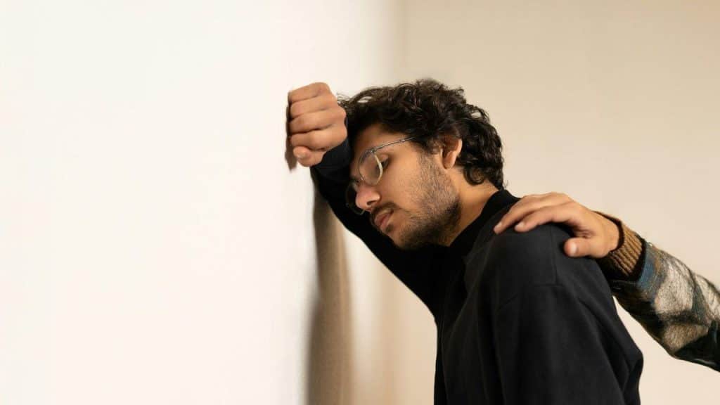 A man leans his head against a white wall while a hand rests on his shoulder.