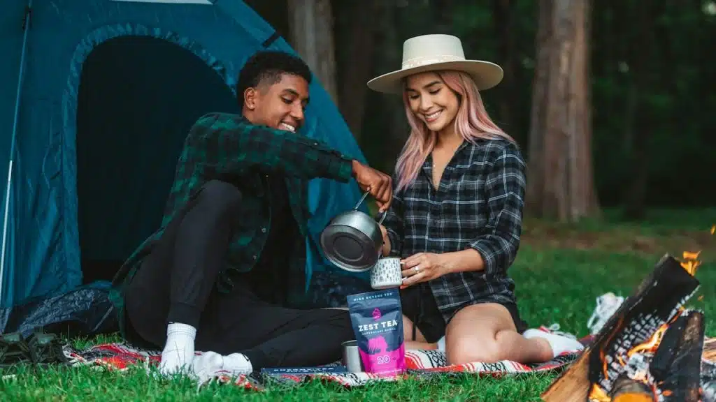 A man pouring tea in the cup of his girlfriend while they are sitting in the grass during camping.