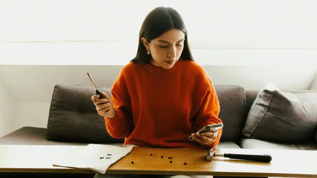 A woman assembling small parts while looking at her phone at a table