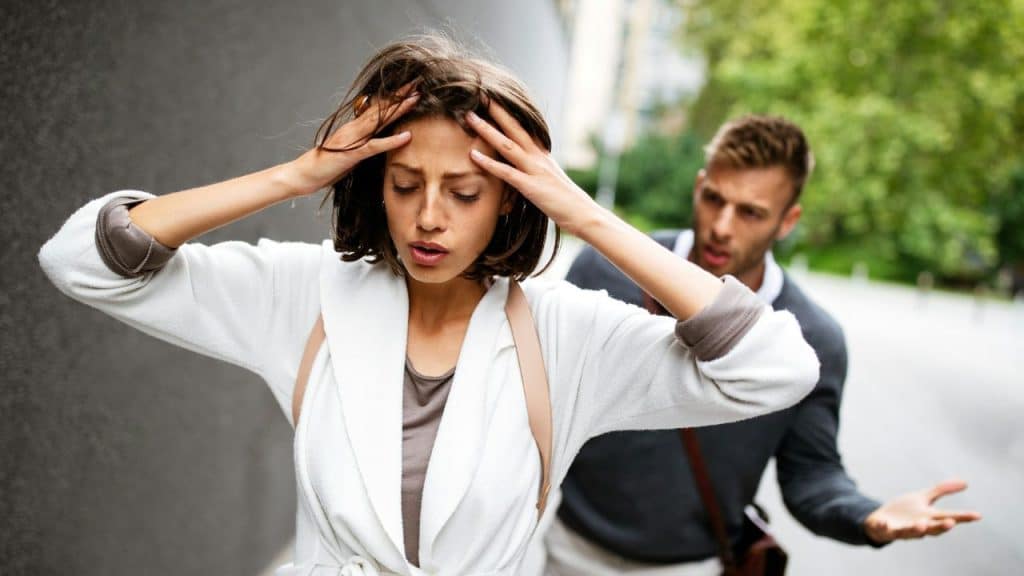 A woman holds her head with both hands while a man gestures behind her outdoors.