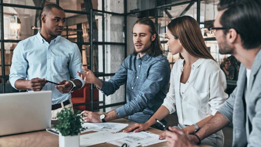 Four colleagues in a modern office stand and sit around a table discussing documents.