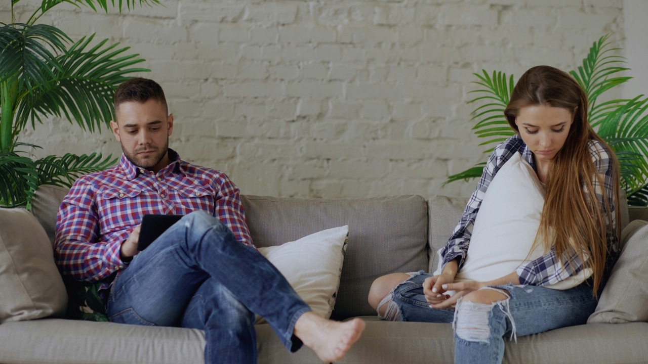 A man and woman sit apart on a sofa, the man using a tablet device.
