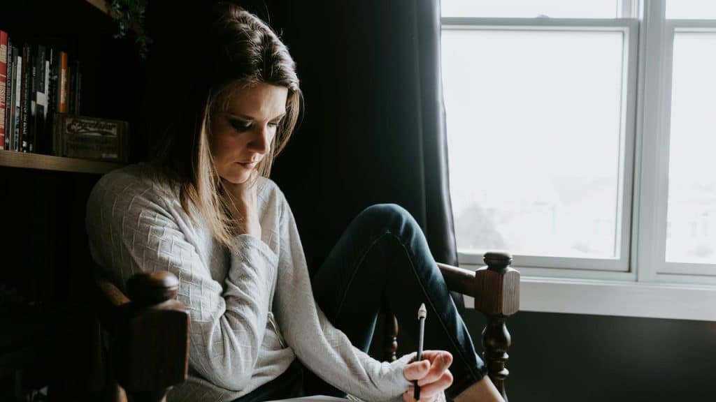 A woman sits by a window writing in a notebook.