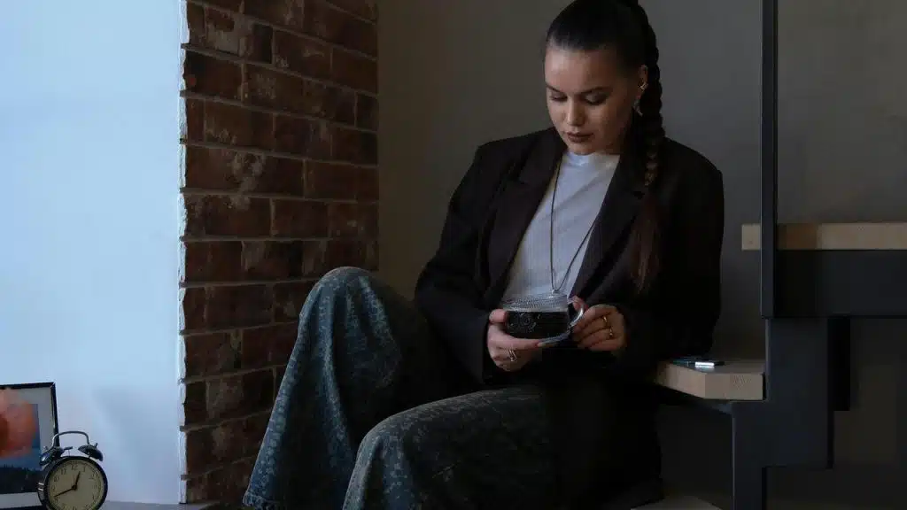 A woman sitting by a window holding a cup and looking down thoughtfully.