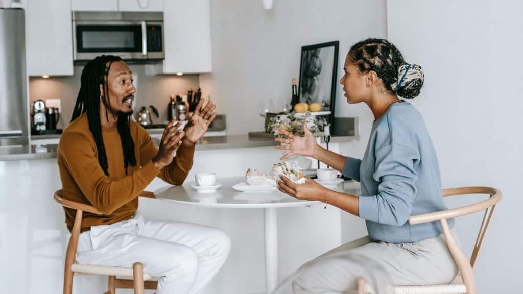 A man and woman sit at a kitchen table talking with their hands raised in gestures.