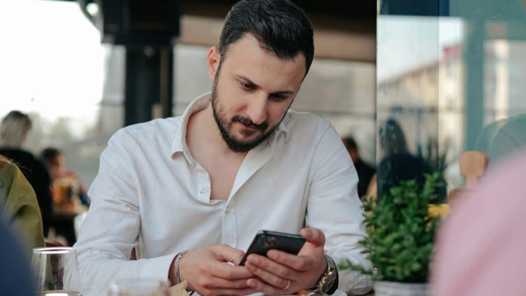 A man with a beard sits at a table while looking down at his smartphone.