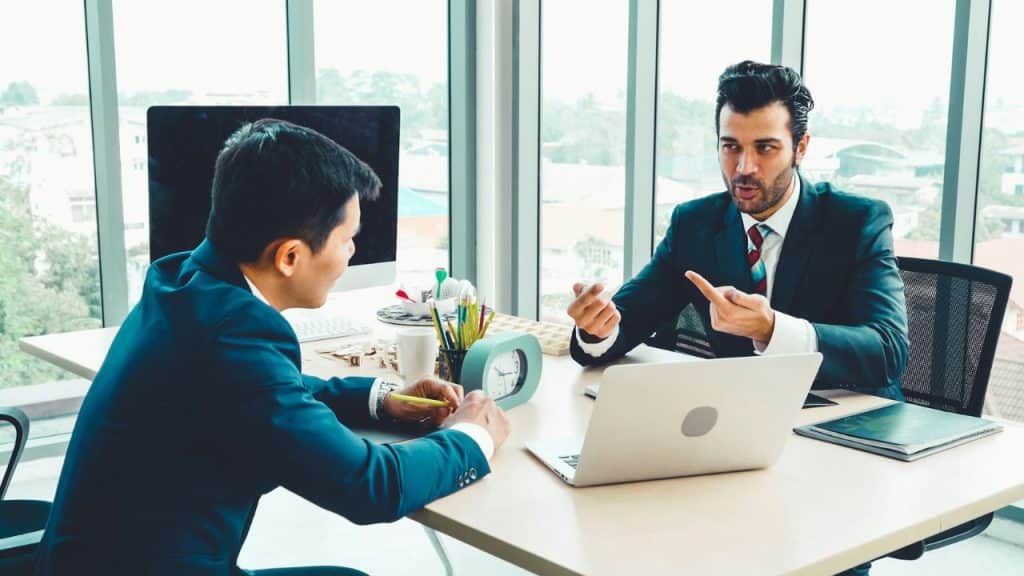 Two men in suits sit at a desk, one gesturing and speaking while the other listens.