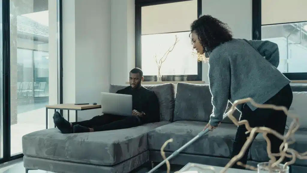 A man uses a laptop on a sofa while a woman vacuums the floor nearby.