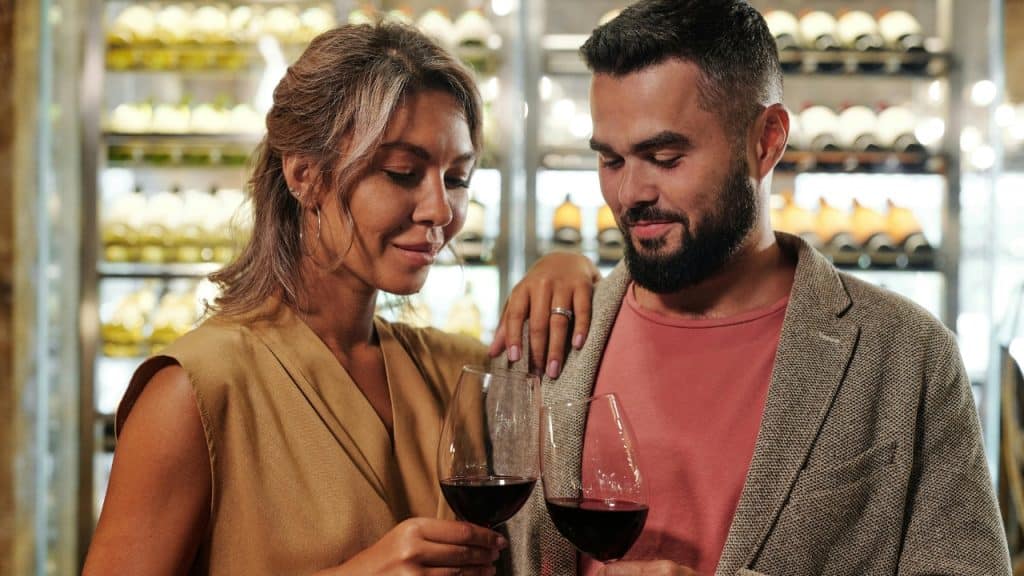 A man and woman stand together in a cellar while holding glasses of red wine.