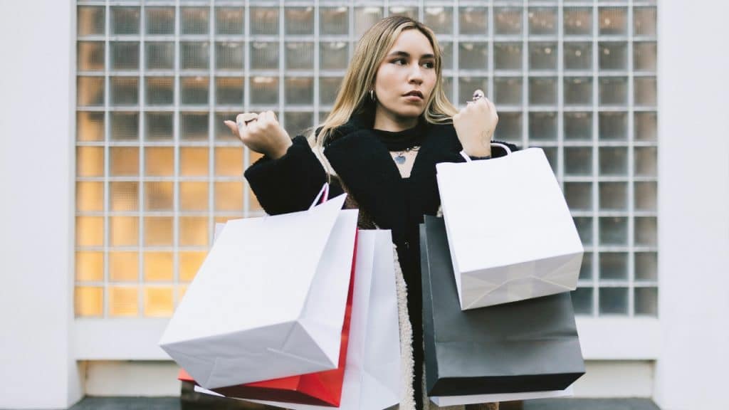 A woman stands in front of a glass block wall holding several white shopping bags.