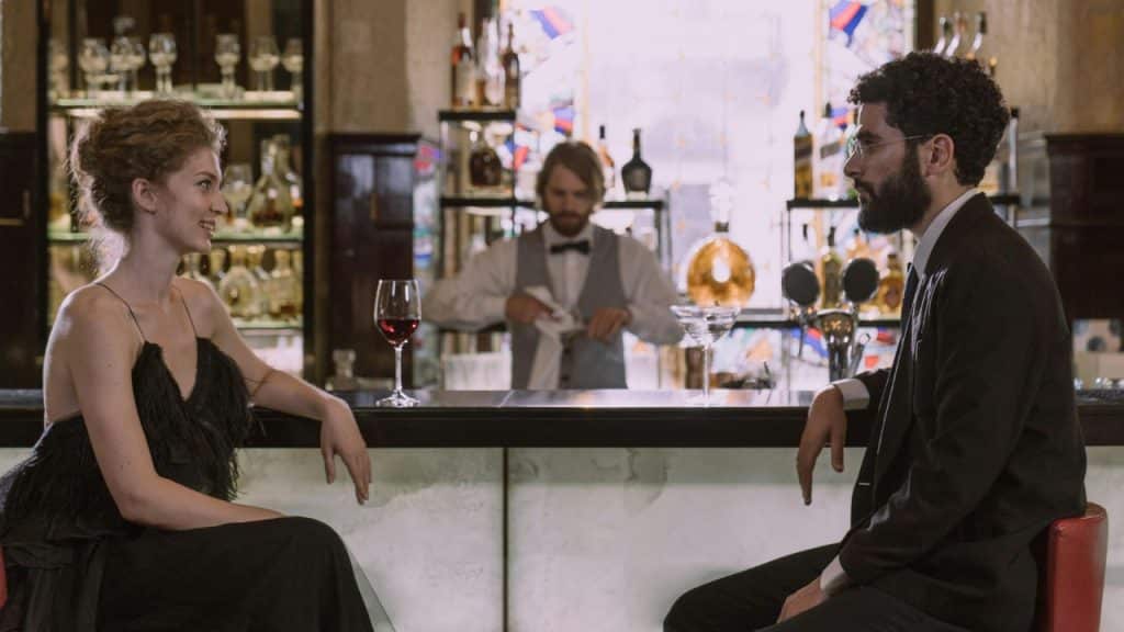 A woman in a black dress and a man in a suit sit at a bar.