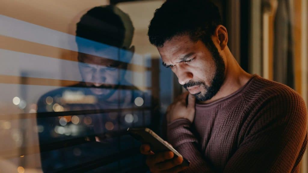 A man looks down at his phone while standing near a window at night