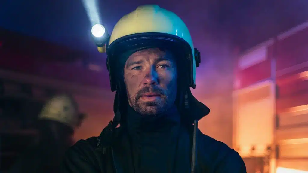 A dirty firefighter man on duty with a fire truck in the background at night looking at the camera.
