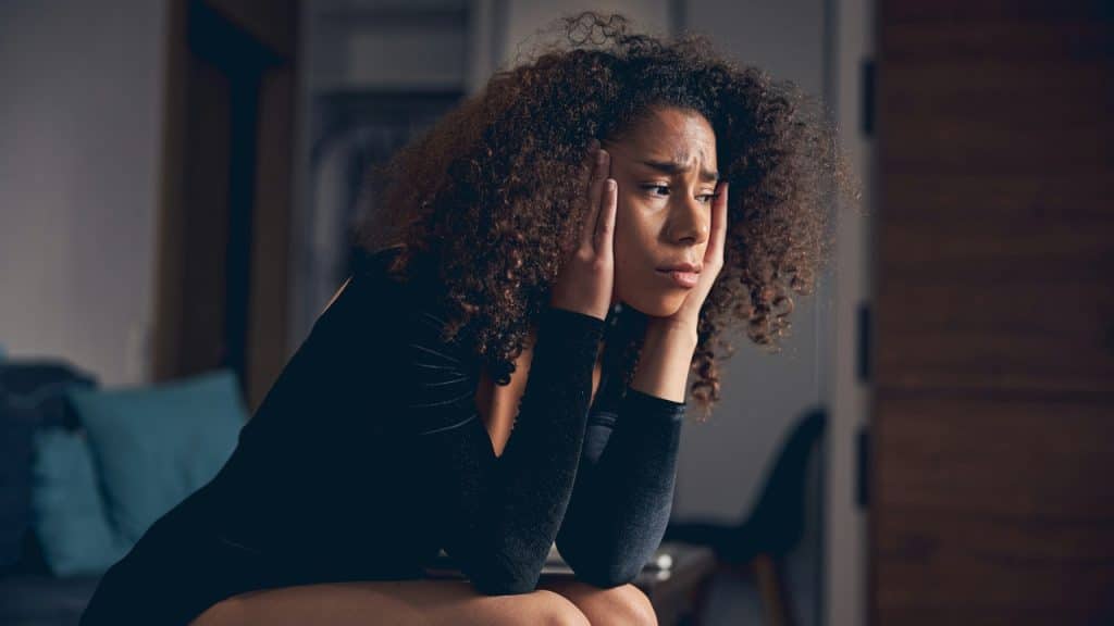 A woman with curly hair sits holding her head with a look of concern.