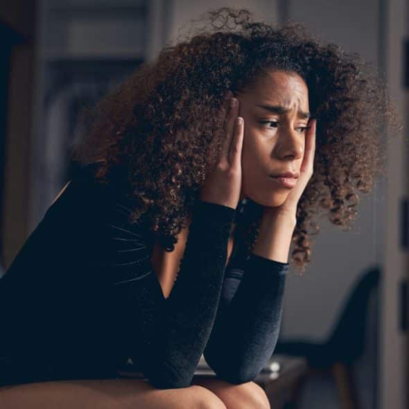 A woman with curly hair sits holding her head with a look of concern.